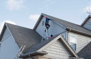 A person clearing snow from a house roof with a roof rake on a sunny winter day.